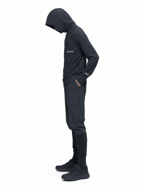 A person in a black hooded jacket and matching black pants stands in profile against a plain white background, with hands in pockets and shoes visible.