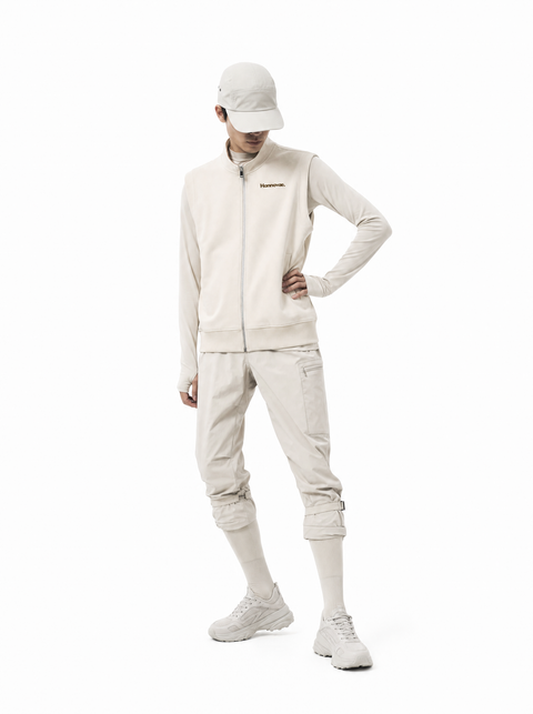 A model wearing a Hannovae light beige outdoor tracksuit consisting of a zip-up jacket, matching pants, a cap, and sneakers, posed against a solid white background.
