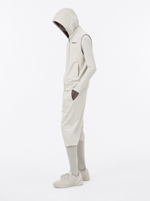 A model wearing a light beige Hannovae hooded vest over a matching long-sleeve top, shorts, and sneakers, shown in profile against a plain white studio background.