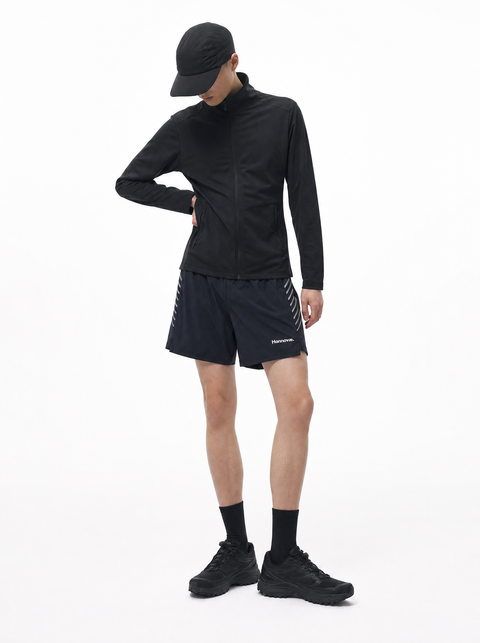 A model wearing a black long-sleeve athletic jacket, black running shorts with white side stripes, a black cap, black socks, and black trail running shoes, photographed against a solid white studio background.